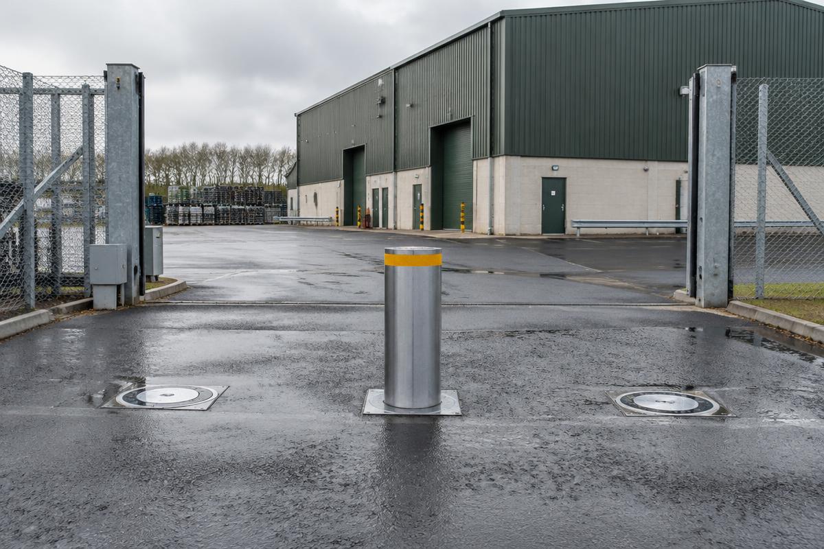 Three automatic rising bollards across a commercial yard entrance, one raised and two lowered flush with the tarmac